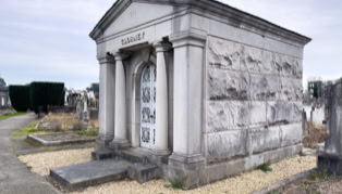 A stone mausoleum with columns and a decorative door, surrounded by a gravel path and other gravestones in a cemetery setting.