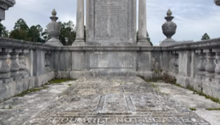A stone tomb with intricate carvings, surrounded by a stone balustrade, with a large inscribed monument in the background.