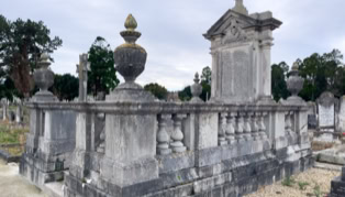A large, ornate stone tomb with decorative urns and a cross on top, surrounded by a stone balustrade in a cemetery setting.