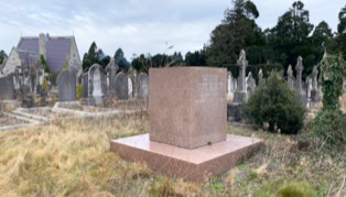 A cemetery with a large, square stone monument in the foreground, surrounded by grass and other gravestones in the background.