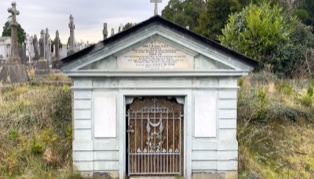 A small mausoleum with a gated entrance, surrounded by grass and other tombstones in the background.
