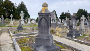 A cemetery with various stone gravestones and crosses, surrounded by grass and pathways under a cloudy sky.