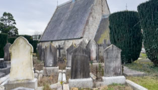 A small stone church with a steep roof surrounded by old gravestones and trimmed hedges.
