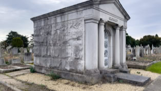 A stone mausoleum with columns and a decorative door, surrounded by a cemetery with gravestones, under a cloudy sky.
