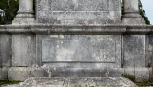 Stone monument with weathered surface, flanked by two columns, surrounded by greenery.