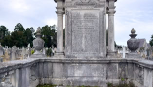 A large, ornate stone tombstone with engraved text, surrounded by a low stone wall, in a cemetery with other gravestones and trees in the background.