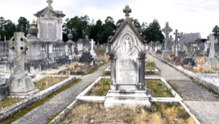A cemetery with multiple gravestones and monuments, featuring a prominent stone cross in the foreground, surrounded by pathways and overgrown grass.