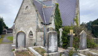 A small stone church with a steep roof, surrounded by old gravestones and overgrown vegetation.