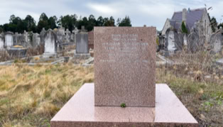 A cemetery with a prominent pink granite tombstone in the foreground, surrounded by grass and other gravestones in the background.