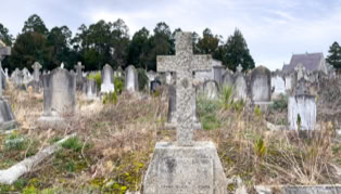 A cemetery with numerous weathered tombstones and crosses, surrounded by overgrown grass and vegetation, under a cloudy sky.