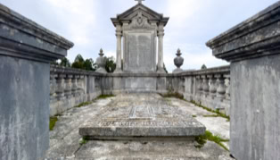 Stone mausoleum with ornate carvings and a cross on top, surrounded by a low stone wall and balustrades, with a cloudy sky in the background.