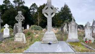A cemetery with several gravestones, including a prominent Celtic cross in the center, surrounded by grass and trees in the background.