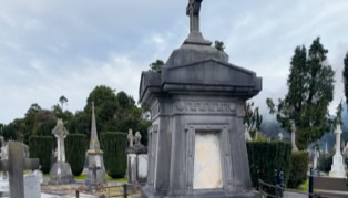 A cemetery with a large stone mausoleum topped by a cross in the foreground, surrounded by various tombstones and trees under a cloudy sky.
