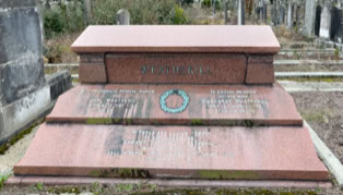 A red granite tomb with inscriptions and a decorative wreath, surrounded by other graves in a cemetery.