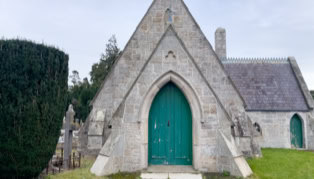 A small stone chapel with a pointed arch doorway painted green, surrounded by grass and trees.
