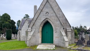 A small stone chapel with a pointed arch and a green door, surrounded by grass and gravestones.
