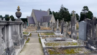 A cemetery with rows of gravestones and a stone pathway leading to a small stone building in the background, surrounded by trees.