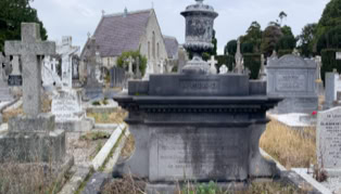 A cemetery with various tombstones and a large stone monument in the foreground, surrounded by overgrown grass, with a chapel in the background.