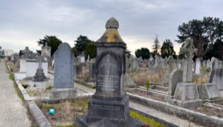 A cemetery with various tombstones and grave markers, including a prominent dark stone monument in the foreground, surrounded by grass and pathways under a cloudy sky.