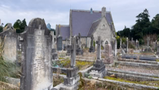 A cemetery with numerous old gravestones and a small stone chapel in the background, surrounded by trees and overcast skies.