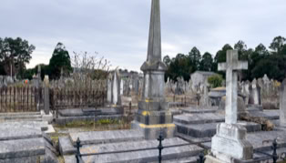 A cemetery with various tombstones, including a tall obelisk and a cross, surrounded by iron fencing and trees in the background under a cloudy sky.