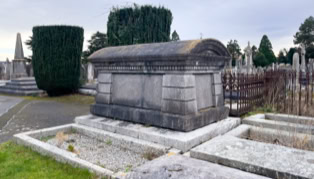 A stone tomb in a cemetery surrounded by other graves and trimmed hedges under a cloudy sky.