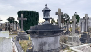 A cemetery with various tombstones and crosses, including a prominent stone monument with an urn on top, surrounded by grass and a cloudy sky.