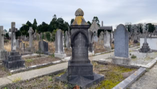 A cemetery with various tombstones and grave markers, including a prominent dark stone monument in the foreground, surrounded by pathways and grass.