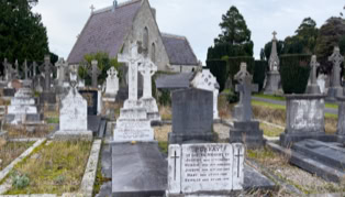 Gravestones in a cemetery with a stone church in the background, surrounded by trees.