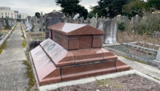 A red granite tomb in a cemetery, surrounded by other gravestones and pathways, with trees and a cloudy sky in the background.