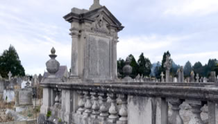 A stone cemetery monument with a cross on top, surrounded by a balustrade, with other gravestones and trees in the background.