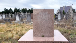 A cemetery with a prominent pink granite tombstone in the foreground, surrounded by grass and other tombstones in the background.